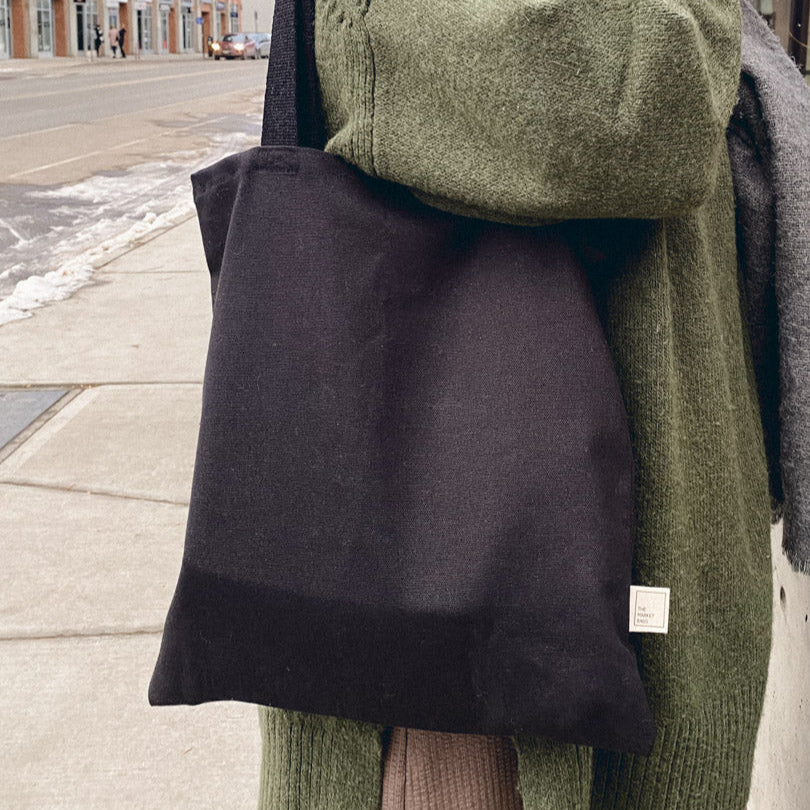 Person wearing a green sweater and holding a black tote bag on a city street.